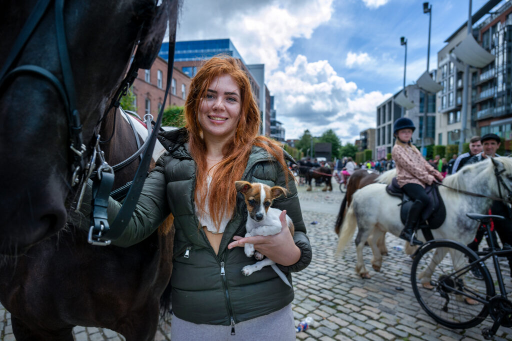 A smiling young red-haired girl with a puppy in her hand holds onto the reins of a horse in Smithfield, Dublin.